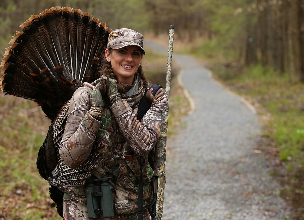 a female hunter carrying a turkey after hunt and also a hunting gun on her shoulder